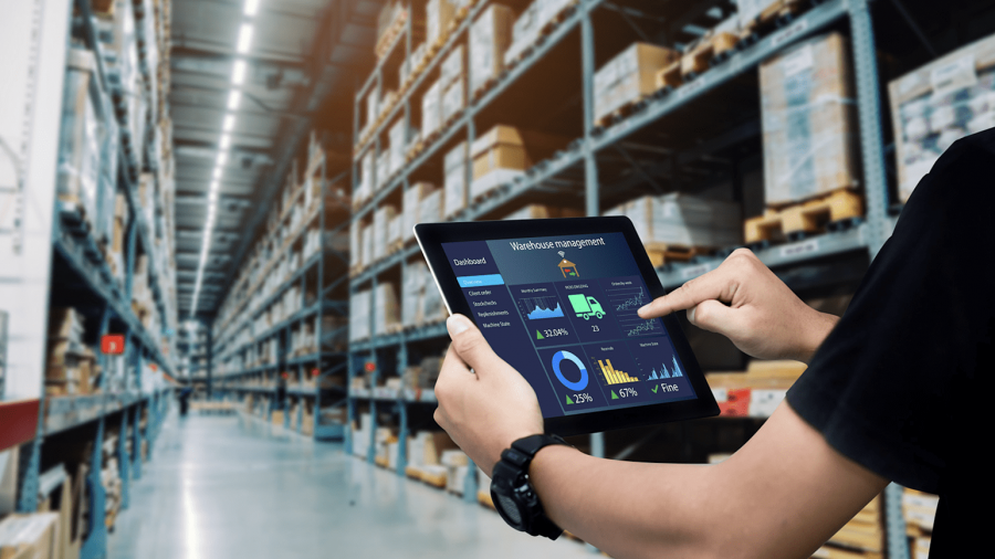 Person using a tablet displaying warehouse automation technologies while standing in a large warehouse with tall shelves filled with boxes and packages.