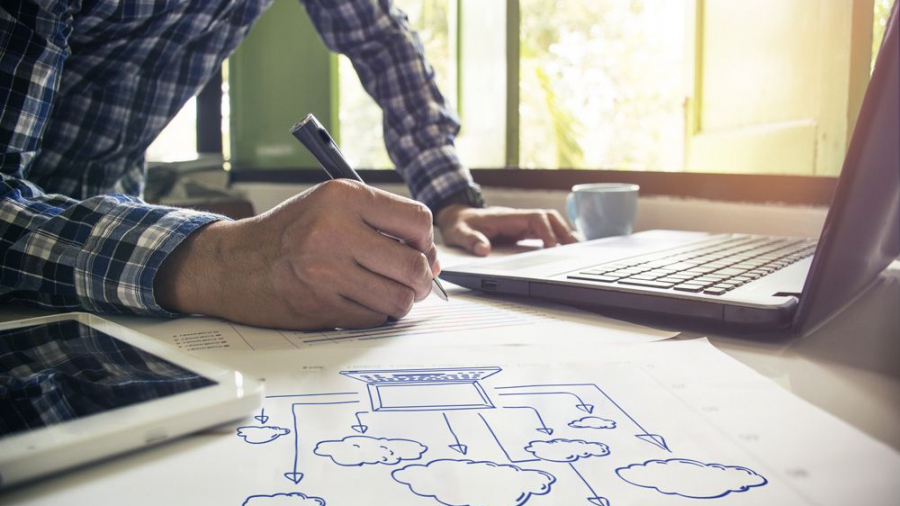 A person writing on paper with cloud diagrams, next to a laptop and tablet running Dynamics 365 Business Central, in a bright office with large windows.