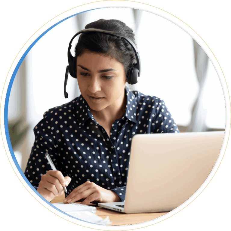 Woman wearing a headset and polka dot shirt sits at a desk, writing in a notebook while looking at a laptop screen, appearing focused and engaged in a Dynamics 365 Customer Service virtual meeting.