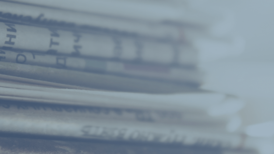 A close-up of a stack of newspapers, viewed from the side, with text visible on the pages. The image has a blue tint and a blurred background.