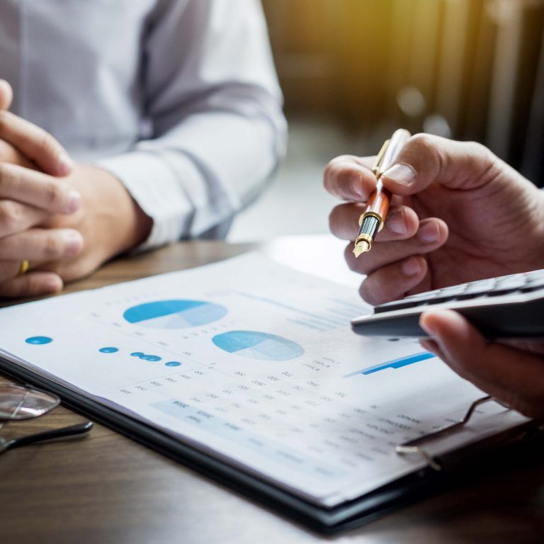 Two people sit at a desk; one holds a pen and calculator, the other has clasped hands. A clipboard with financial charts, glasses, and an enVista 365 pricing sheet are on the table, suggesting a discussion about finances or planning.