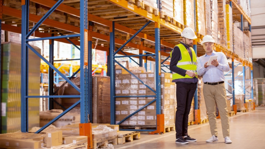 Two men wearing hard hats stand in a warehouse beside tall shelves stacked with boxes. One man in a safety vest and the other with a clipboard discuss inventory, possibly reviewing data from their labor management system.