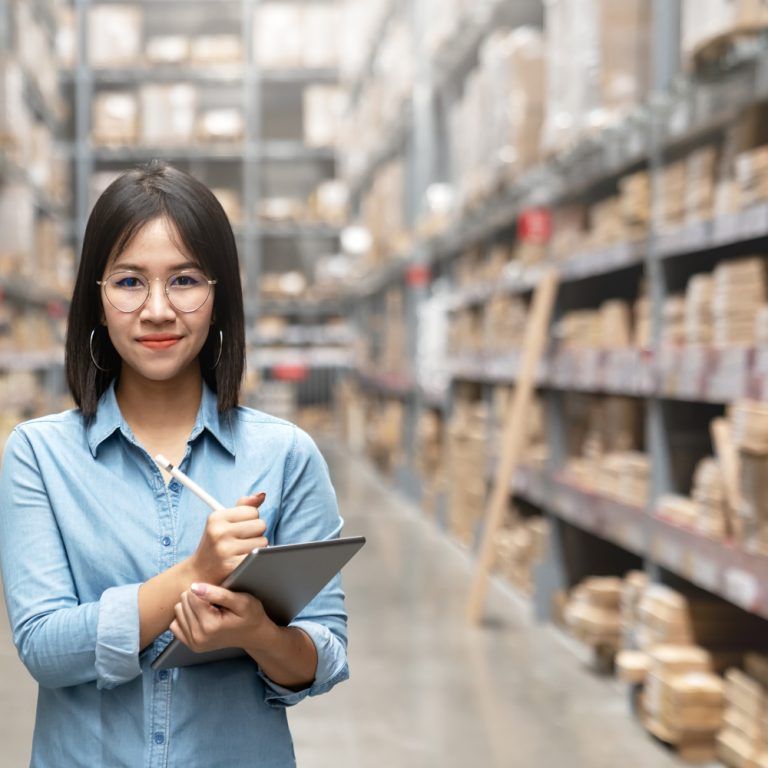 A woman with glasses and a blue shirt holds a clipboard and pen, standing in a warehouse aisle lined with boxes. She smiles confidently at the camera, ready to manage inventory efficiently with Dynamics 365 Business Central.