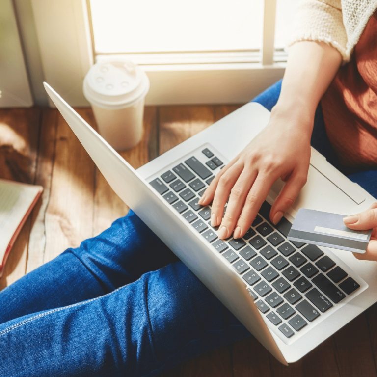 A person sitting on the floor uses a laptop while holding a credit card, with a notebook, pen, coffee cup, and potted plant nearby, suggesting online shopping or banking at home.
