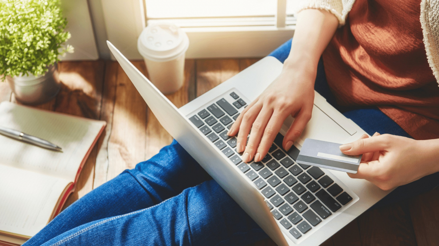 A person sitting on the floor uses a laptop while holding a credit card, with a notebook, pen, coffee cup, and potted plant nearby, suggesting online shopping or banking at home.