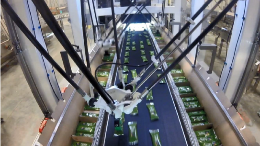 Overhead view of an automated robotic arm in a factory sorting green packaged products on a conveyor belt, surrounded by machinery and industrial equipment.