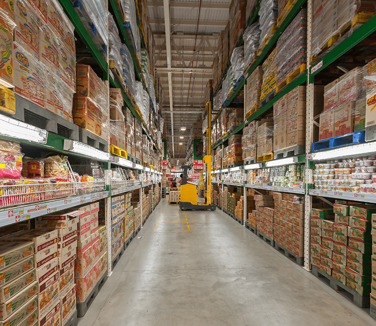 A wide aisle in a warehouse-style grocery store features tall shelves stacked with packaged food products, highlighting the importance of 3PL selection for efficient logistics, with a yellow pallet jack parked at the end of the aisle.