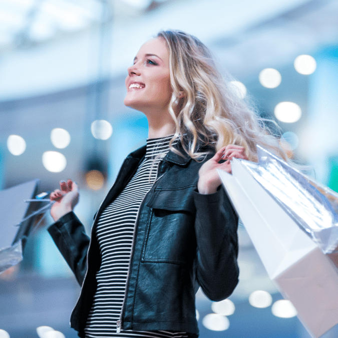 A smiling woman with long blonde hair wearing a striped top and black leather jacket holds several shopping bags in a brightly lit mall.