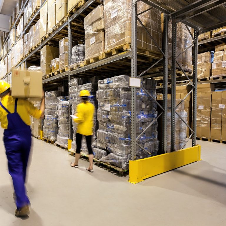 Two workers wearing yellow helmets and uniforms move boxes in a brightly lit warehouse filled with shelves stacked with pallets of goods and cardboard boxes.