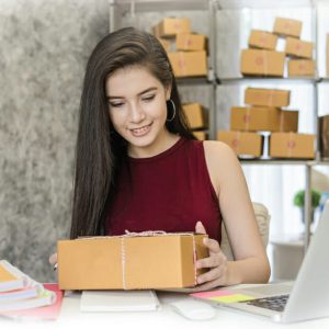 A young woman with long dark hair sits at a desk, smiling as she holds a brown package. Behind her are shelves filled with similar boxes, highlighting her online business success powered by smart technology solutions in her home office.