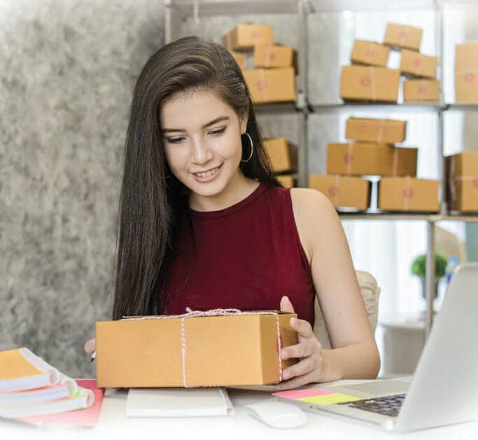 A young woman with long dark hair sits at a desk, smiling as she holds a brown package. Behind her are shelves filled with similar boxes, highlighting her online business success powered by smart technology solutions in her home office.