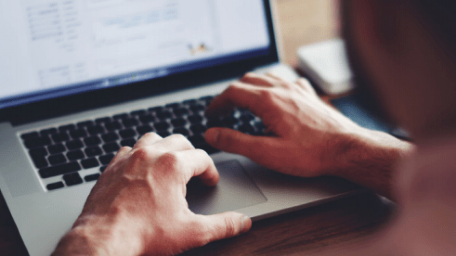 A person uses a laptop at a wooden desk, typing with both hands. The screen displays documents or spreadsheets, and a smartphone lies nearby. The focus is on the hands and laptop keyboard.