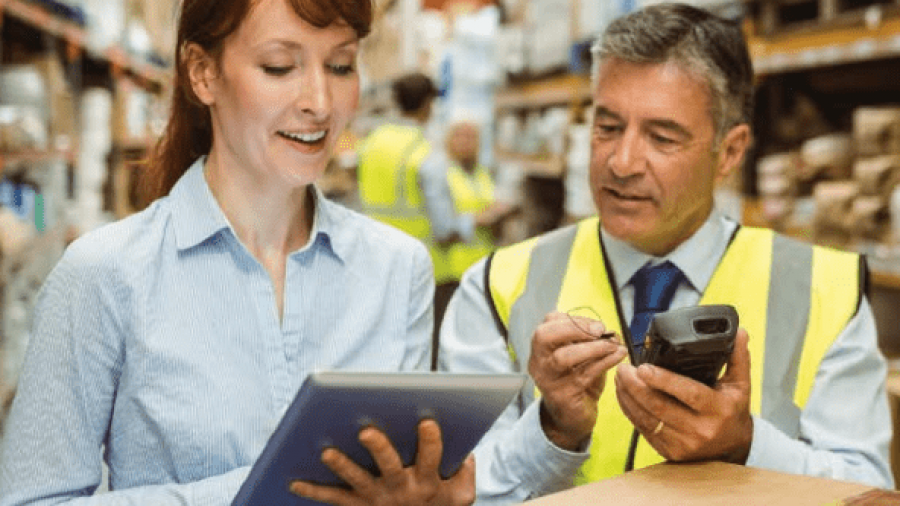 A woman holding a tablet and a man in a safety vest using a handheld device are talking in a warehouse filled with shelves and boxes.