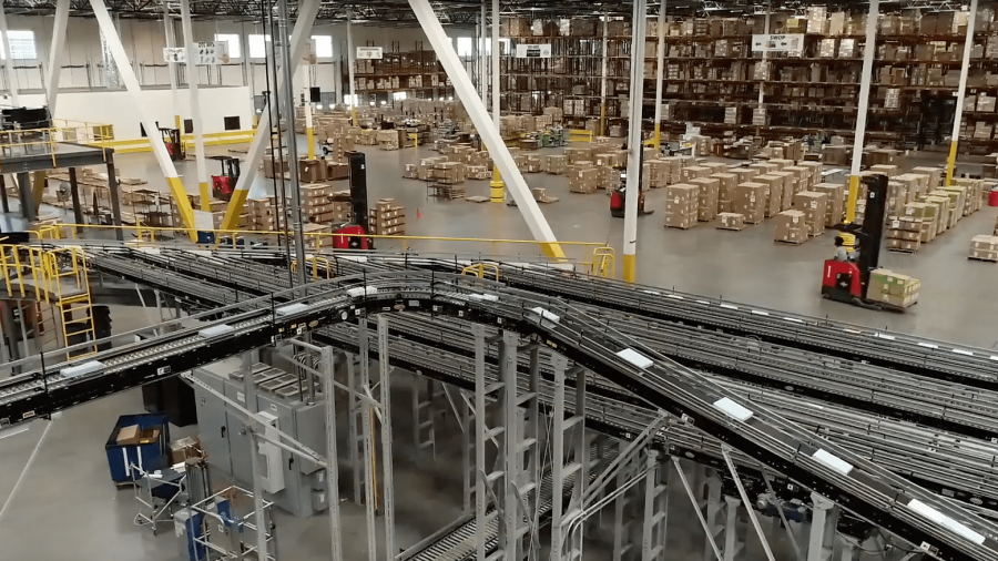 Large warehouse interior with tall shelves stacked with boxes, automated conveyor belts in the foreground, and several forklifts moving pallets around the spacious floor.