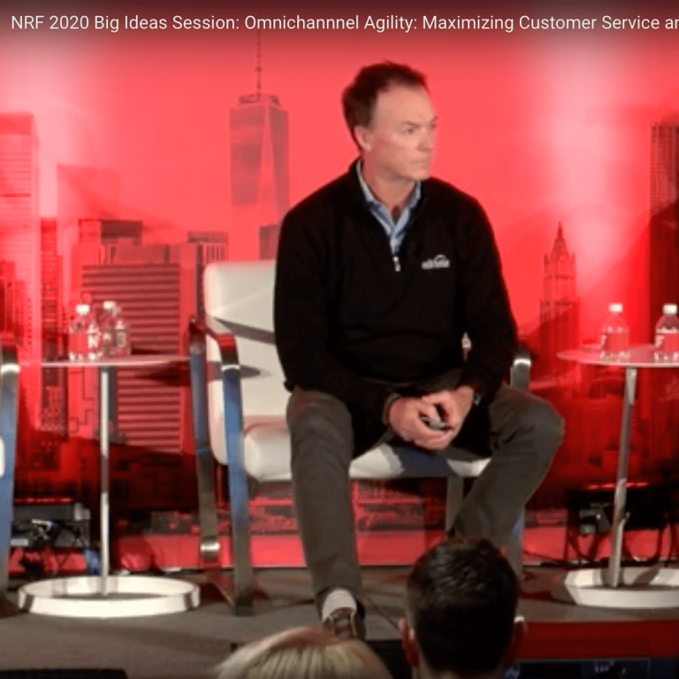 A man in a dark blazer and gray pants sits on a stage with a red backdrop featuring a city skyline graphic, speaking at the NRF 2020 Big Ideas Session. A small table with bottled water is beside him.