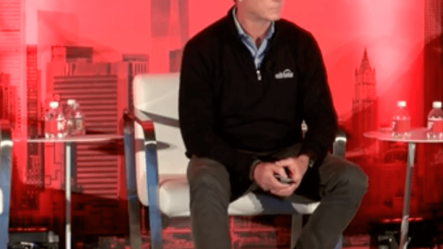 A man in a dark blazer and gray pants sits on a stage with a red backdrop featuring a city skyline graphic, speaking at the NRF 2020 Big Ideas Session. A small table with bottled water is beside him.