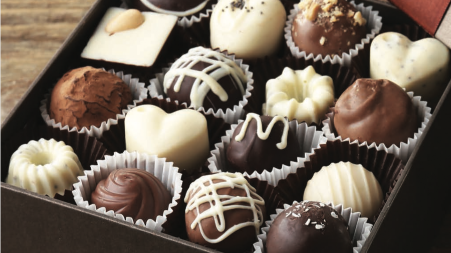 An open box of assorted chocolates in various shapes and colors, including hearts, domes, and squares, with decorative toppings, arranged neatly in paper cups on a wooden surface.