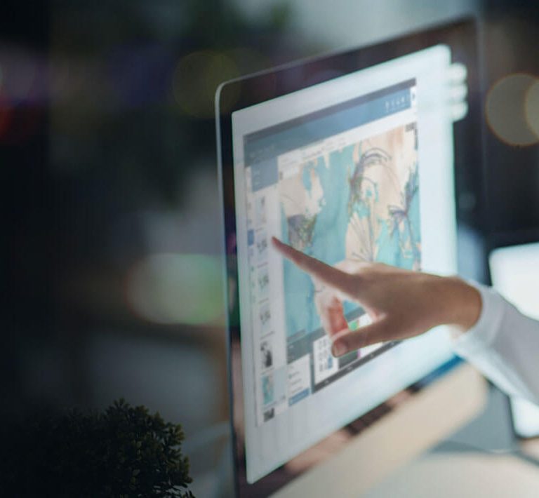 A person wearing glasses talks on the phone while pointing at a computer screen displaying colorful graphics or data in a dimly lit office setting.