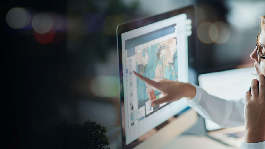 A person wearing glasses talks on the phone while pointing at a computer screen displaying colorful graphics or data in a dimly lit office setting.