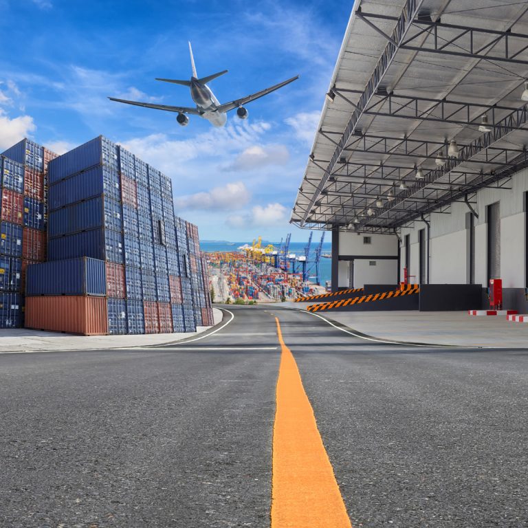 A commercial airplane flies over a shipping port with stacked cargo containers and a warehouse loading dock under a partly cloudy sky.