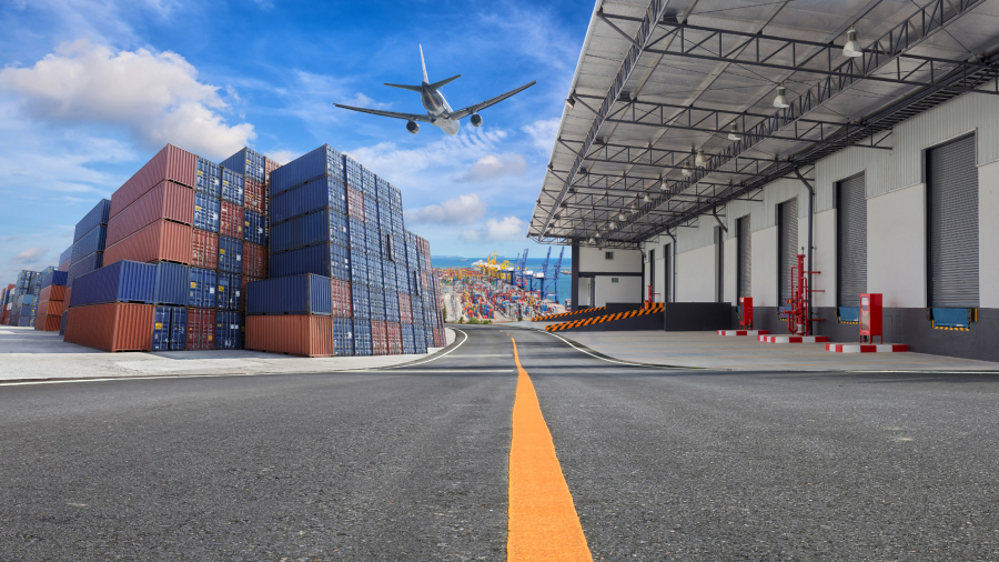 A commercial airplane flies over a shipping port with stacked cargo containers and a warehouse loading dock under a partly cloudy sky.