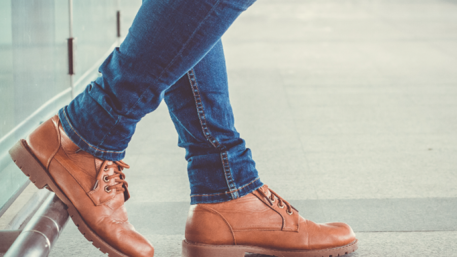 A person wearing blue jeans and tan leather boots stands indoors, leaning against a glass wall with one foot crossed over the other on a tiled floor. The background is out of focus.