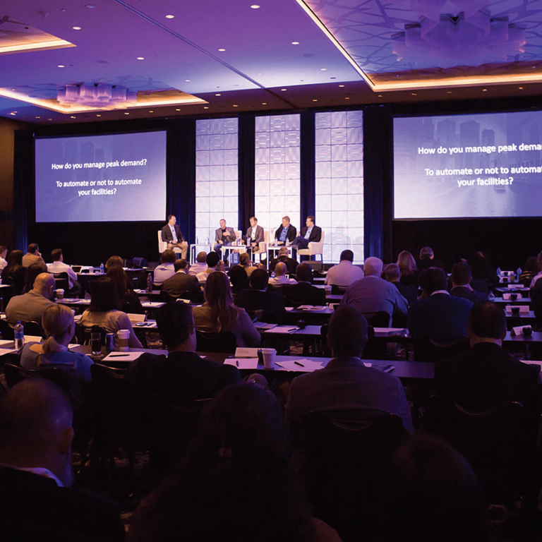 A conference room filled with attendees faces a stage where four panelists discuss, "How do you manage peak demand? To automate or not to automate your facilities?"—all within the context of a unified commerce strategy.