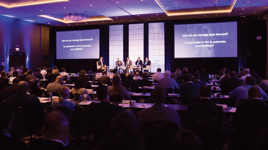 A conference room filled with attendees faces a stage where four panelists discuss, "How do you manage peak demand? To automate or not to automate your facilities?"—all within the context of a unified commerce strategy.