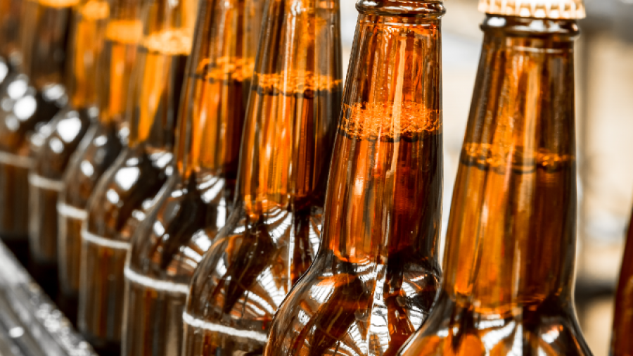 A row of brown glass beer bottles with caps moves along a conveyor belt in a brewery, reflecting bright factory lights.