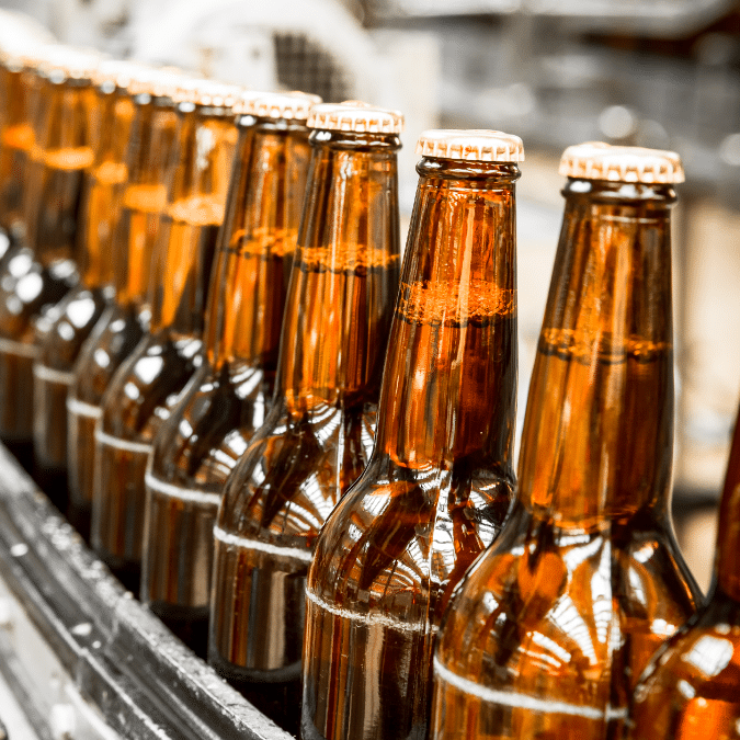 A row of brown glass beer bottles with caps moves along a conveyor belt in a brewery, reflecting bright factory lights.