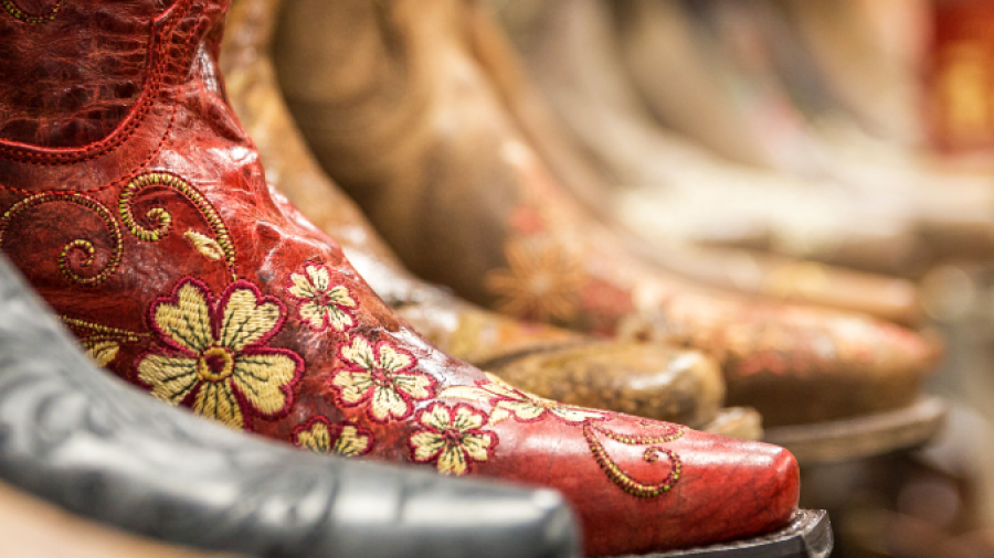A close-up of a bright red cowboy boot with yellow floral embroidery, displayed among other brown and tan cowboy boots on a shelf in a store.