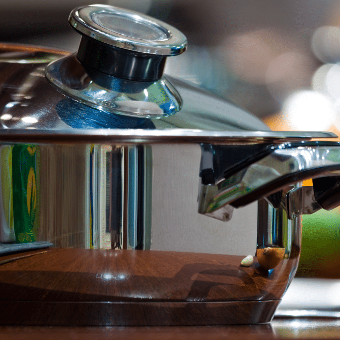 A close-up of a shiny stainless steel pot with a glass lid on a wooden surface. The pots handle and lid knob are visible, and reflections can be seen on the metal surface.