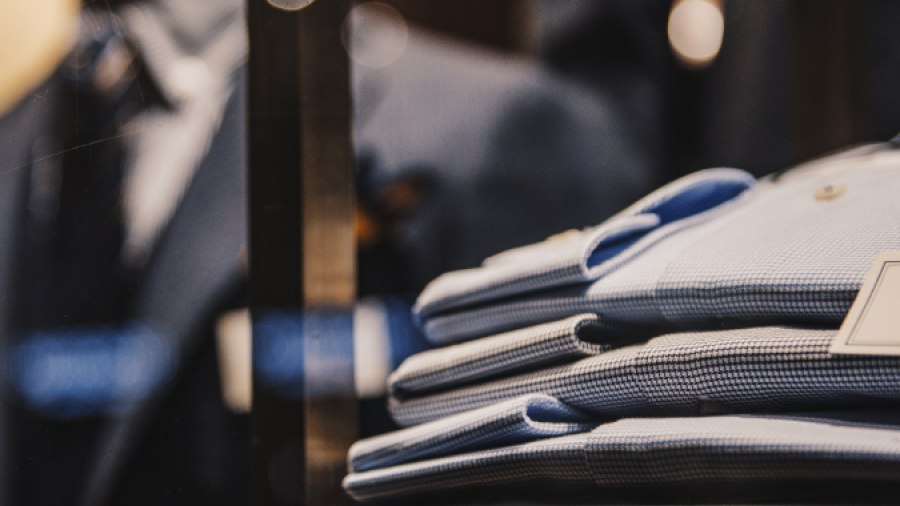 Folded blue dress shirts neatly stacked on a shelf in a clothing store, with blurred suits hanging in the background and warm lighting creating a stylish, elegant atmosphere.