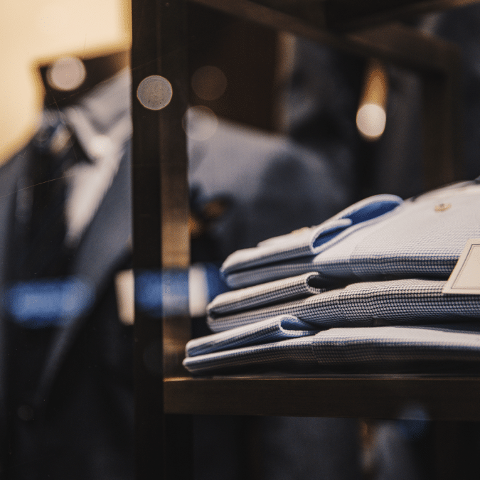 Folded blue dress shirts neatly stacked on a shelf in a clothing store, with blurred suits hanging in the background and warm lighting creating a stylish, elegant atmosphere.