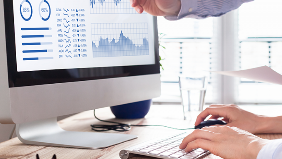 Two people work at a desk with a computer displaying charts and graphs. One person points at the screen, while the other types on the keyboard. Papers, glasses, and a glass of water are on the desk.