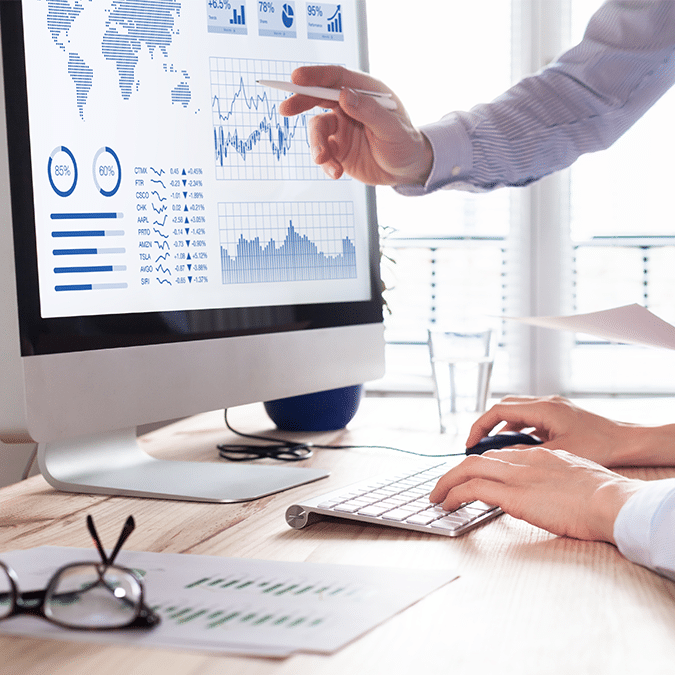 Two people work at a desk with a computer displaying charts and graphs. One person points at the screen, while the other types on the keyboard. Papers, glasses, and a glass of water are on the desk.