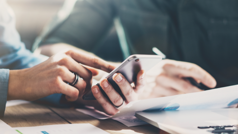 Two people sit at a desk with documents and a notebook, one person holding a smartphone while the other points at the screen, suggesting collaboration or discussion.