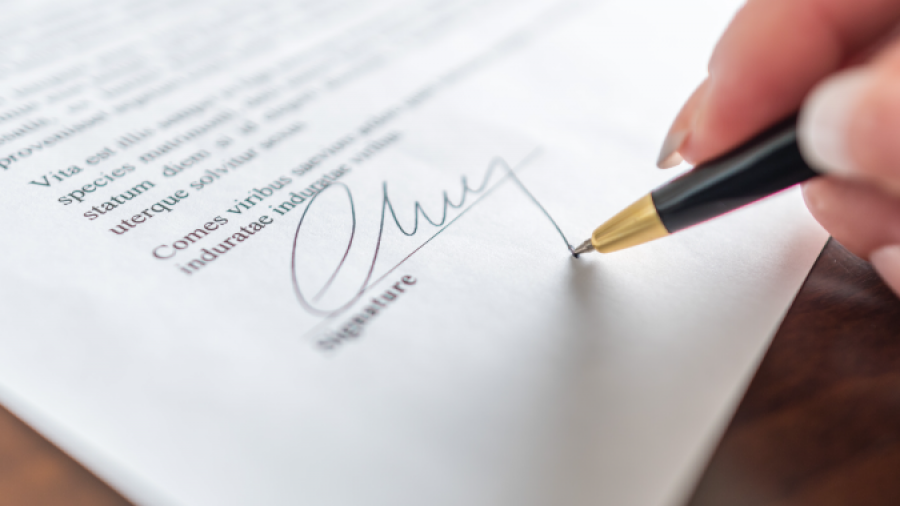 A close-up of a hand holding a pen and signing a document on a wooden surface. The signature is being written on a printed contract or agreement.