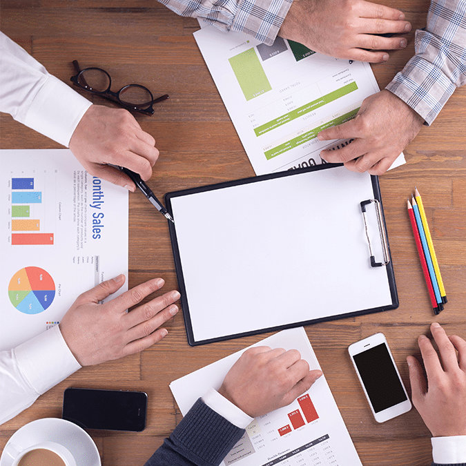 Four people sit around a wooden table with charts, graphs, a clipboard, pencils, glasses, a phone, and coffee, discussing business or sales data in a meeting.