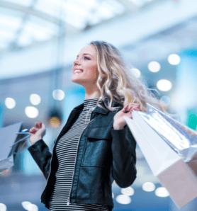 A smiling woman with long blonde hair holds several shopping bags while standing indoors, wearing a black leather jacket and striped shirt. The background is brightly lit and blurred.