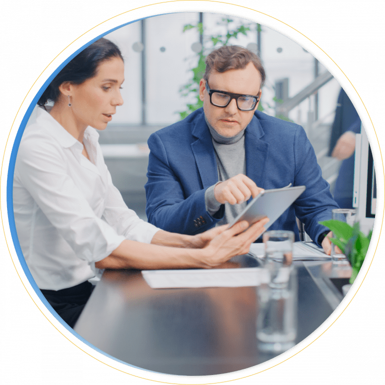 Two professionals sit at a desk in an office, discussing information displayed on a tablet. The man points at the screen while the woman holds the device, reviewing d365 sales data. Papers, a laptop, and a glass of water are on the desk.