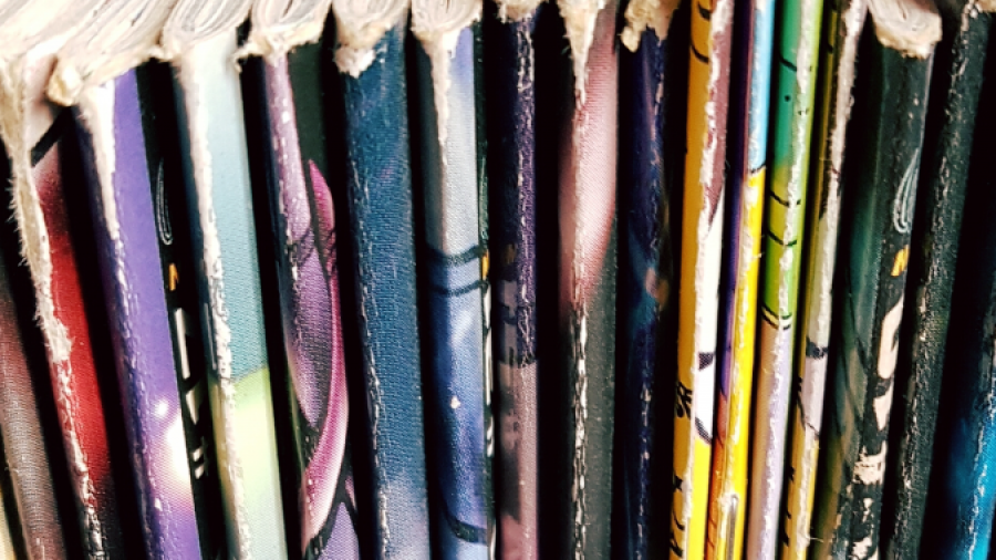 Close-up view of the top edges of several colorful comic books or graphic novels lined up vertically on a shelf, with worn pages and spines visible.