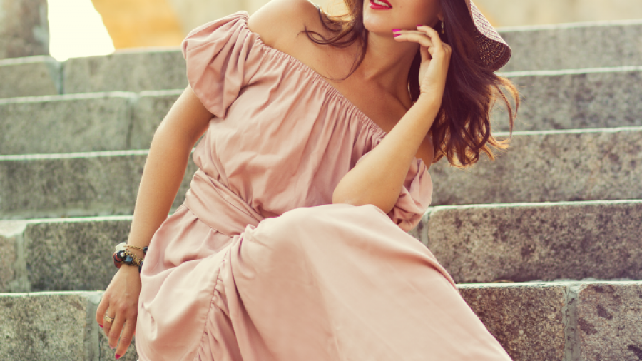 A woman in a flowing pink off-shoulder dress and wide-brimmed hat sits on stone steps, resting her chin on her hand and looking to the side with a relaxed, thoughtful expression.