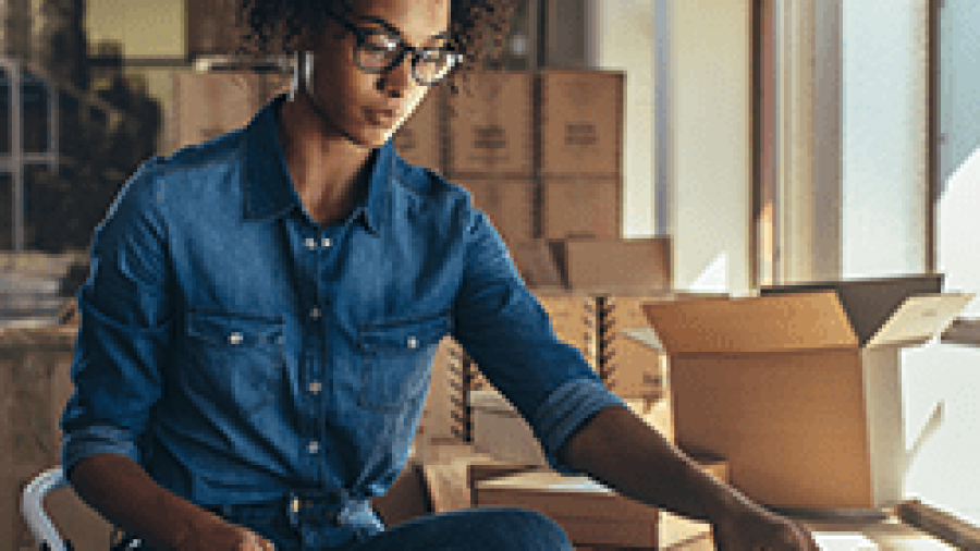 A woman wearing glasses and a denim shirt sits by a window, packing items into a cardboard box in a room filled with more boxes, suggesting a small business or shipping workspace.