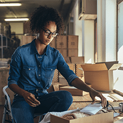 A woman wearing glasses and a denim shirt sits by a window, packing items into a cardboard box in a room filled with more boxes, suggesting a small business or shipping workspace.