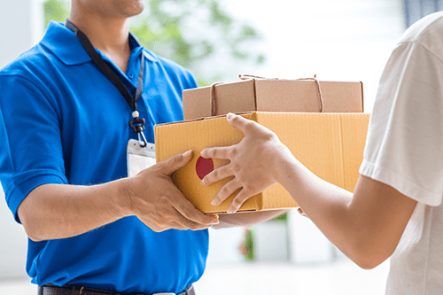 A delivery person in a blue shirt hands multiple packages, including a large yellow box, to another person. Both faces are out of frame, focusing on the exchange of parcels.