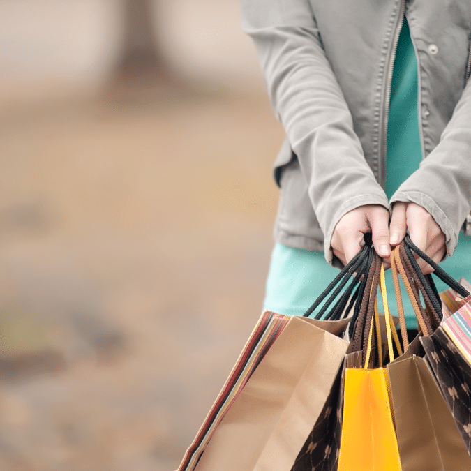 A person wearing a teal dress and gray jacket holds several colorful shopping bags in both hands, standing outdoors with a blurred background.
