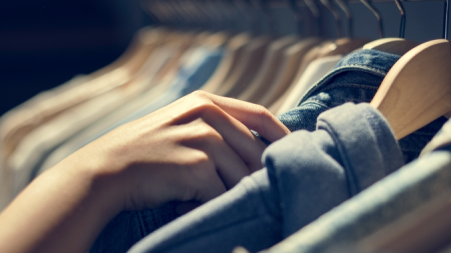 A person’s hand is browsing through a row of clothes on hangers, selecting or inspecting a garment in a softly lit store or closet.