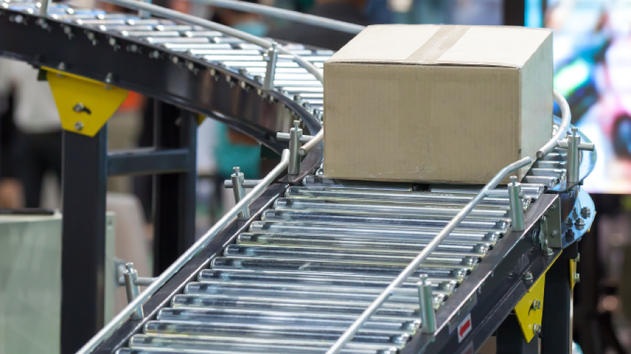 A cardboard box moves along a curved conveyor belt in a warehouse or factory setting, with blurred people and machinery in the background.