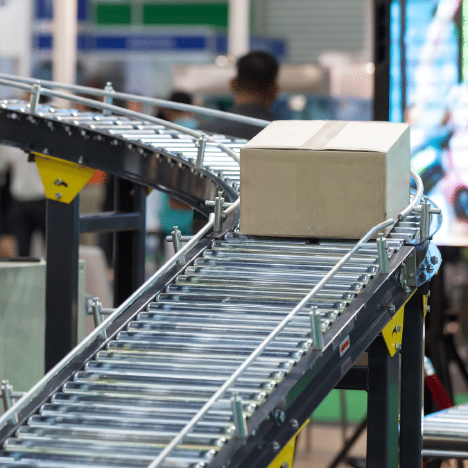 A cardboard box moves along a curved conveyor belt in a warehouse or factory setting, with blurred people and machinery in the background.
